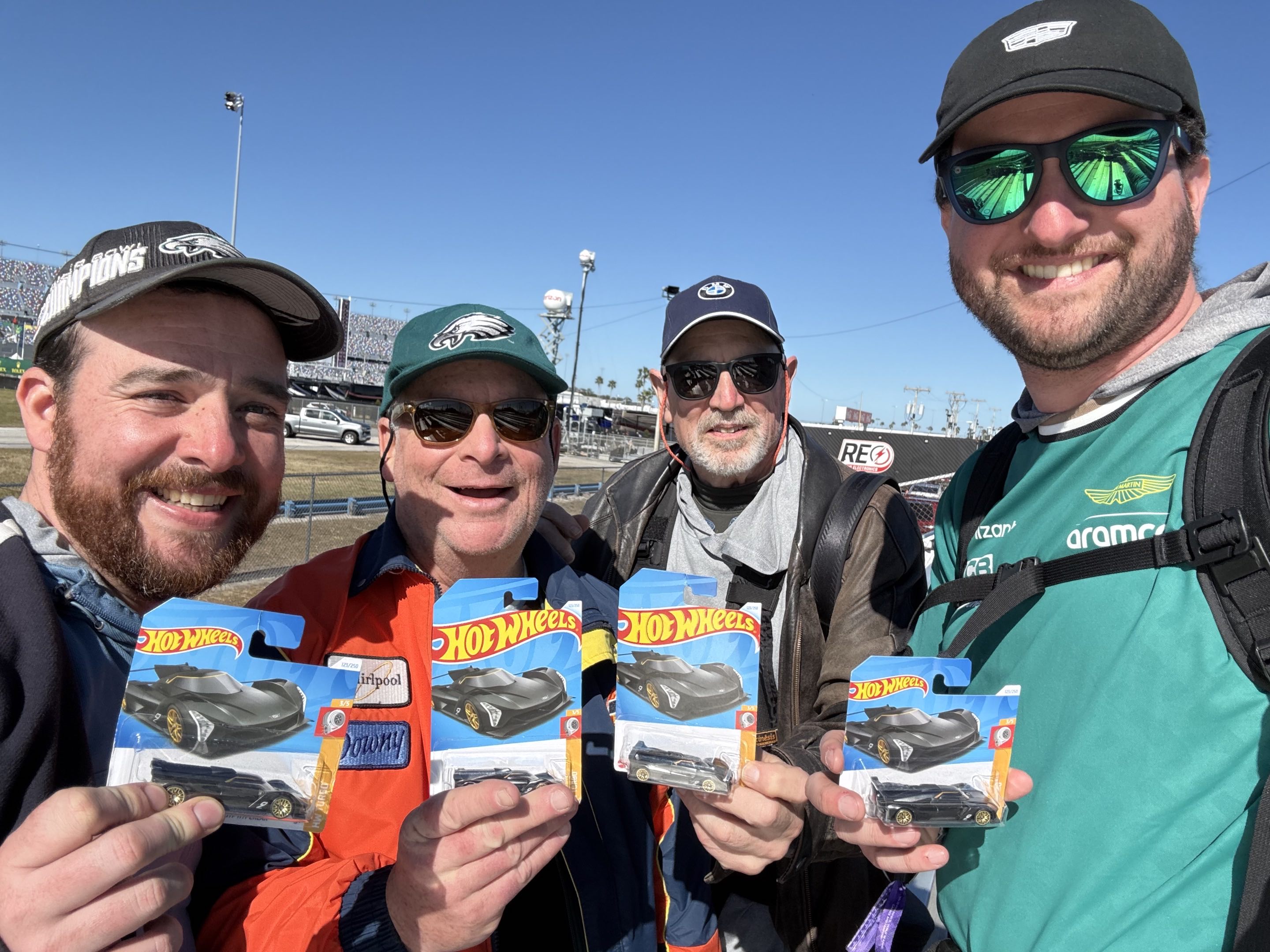 Two thirtysomething men and two of their elders in motorsports fan outfits holding up Hot Wheels packages at a race track